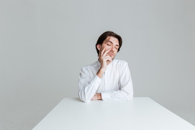 Nadenkende man met gesloten ogen die aan iets denkt terwijl hij aan de tafel zit geïsoleerd op de grijze muur