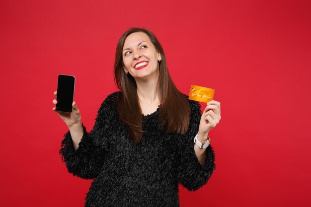 Nadenkende vrouw in bont trui opzoeken, met mobiele telefoon met lege lege scherm creditcard geïsoleerd op rode muur achtergrond. Mensen oprechte emoties, lifestyle concept. Bespotten kopie ruimte.