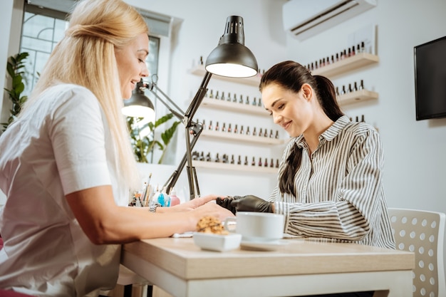 Nail artist. Pleasant dark-haired nail artist feeling good talking to her constant client while working