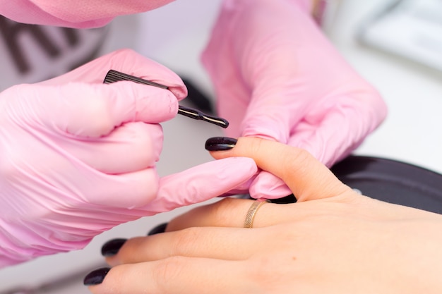 Concetto di cura delle unghie e manicure. le mani del manicure di primo piano nei guanti rosa sta dipingendo lo smalto nero sulle unghie del cliente.