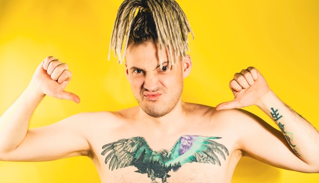 Naked handsome young man 30s with tattoo pointing gesture of\
likes on himself attractive confident guy with blonde dreadlocks\
showing thumbs up on himself in studio on isolated yellow\
background