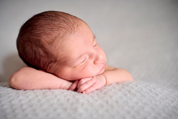 Голый новорожденный спит на руках поверх серого одеяла.
