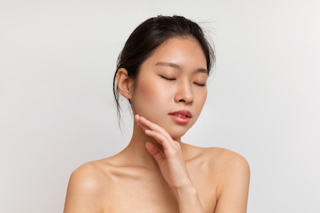 Naked sensual korean lady touching her face and enjoying silky touch after using nourishing cream