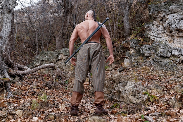 A naked viking with a sheathed sword hanging on his back stands at the foot of the mountain