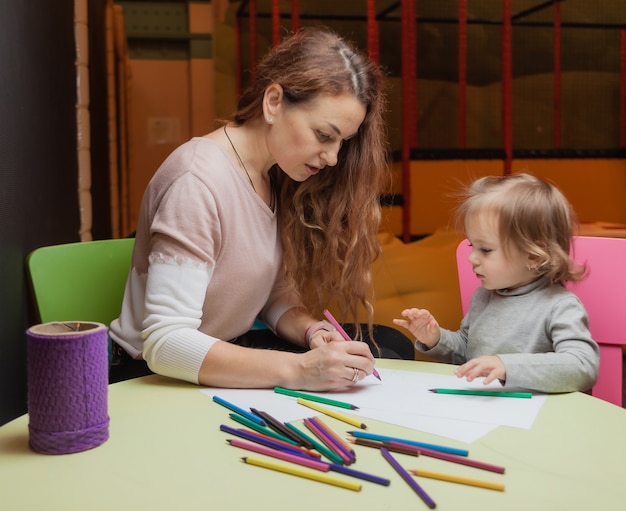 乳母は、子供の娯楽センターのテーブルに座っている間、色鉛筆で描く方法を小さな女の子に教えます