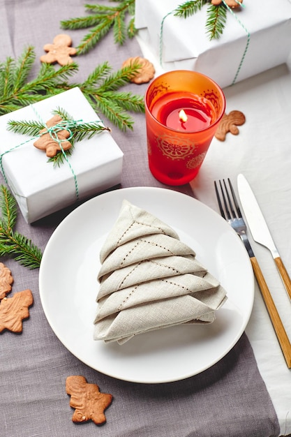 Napkin in the form of a Christmas tree on a plate on white tablecloth with gifts and decorations with fir sprigs and gingerbread cookies
