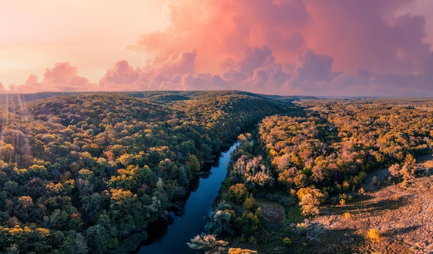 Narrow calm river branches running across autumn landscape with colorful dense forests under cloudy pink sky at sunset aerial view