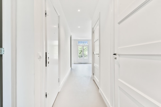 Узкий коридор с дверями, ведущими в светлую пустую комнату с окном в современной квартире