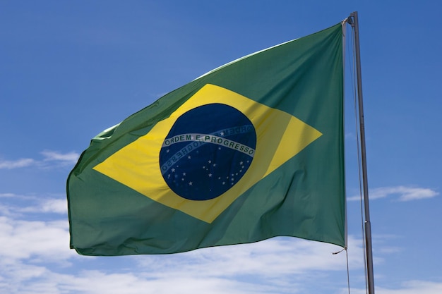 Foto la bandiera nazionale del brasile oscilla nel vento su un albero alto e sullo sfondo di un cielo blu leggermente nuvoloso