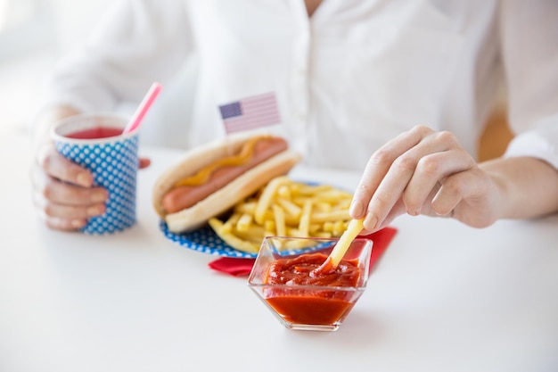 Feste nazionali, celebrazione, cibo e concetto di patriottismo - primo piano di donna che mangia patatine fritte con hot dog e beve succo dal bicchiere di carta al 4 luglio alla festa del giorno dell'indipendenza americana