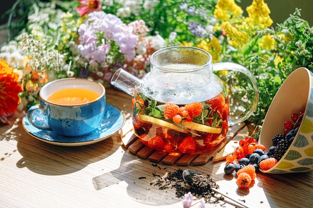 Tè aromatico naturale alla frutta in teiera trasparente e in tazza tra frutti di bosco e fiori tè aromatico riscaldante dal profondo aroma di frutti di bosco e fiori di campo