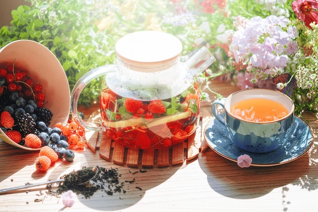 Natural aromatic fruit tea in a transparent teapot and in a cup among berries and flowers warming aromatic tea with a deep aroma of berries and wildflowers