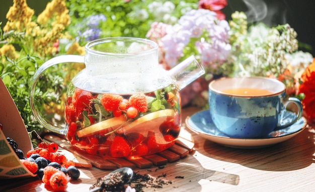 Натуральный ароматный фруктовый чай в прозрачном чайнике и в чашке среди ягод и цветов согревающий ароматный чай с глубоким ароматом ягод и полевых цветов