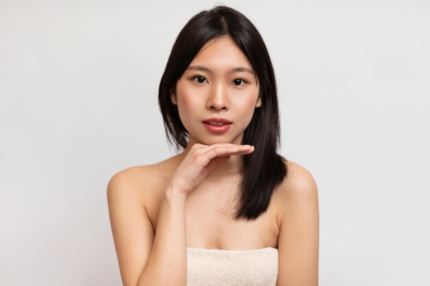 Natural beauty Portrait of young asian lady wrapped in towel touching chin and looking at camera white background