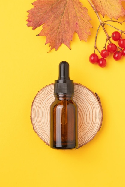 Natural essential oil of viburnum in bottle with a dropper on yellow background with a branch of autumn ripe viburnum