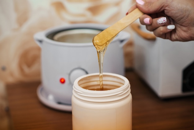 Natural hot sugar paste for depilation in a jar and on a wooden spatula in the hands of a woman beautician