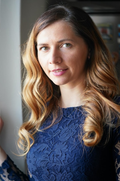 Natural light portrait of young woman with long waves hair, wearing dark blue dress, blurred steel fridge background