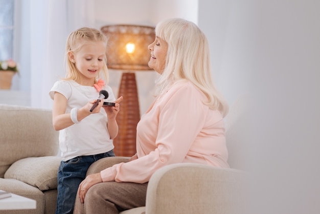 Natural makeup. Pleasant cheerful blonde girl holding powder and looking at it while being together with her grandmother