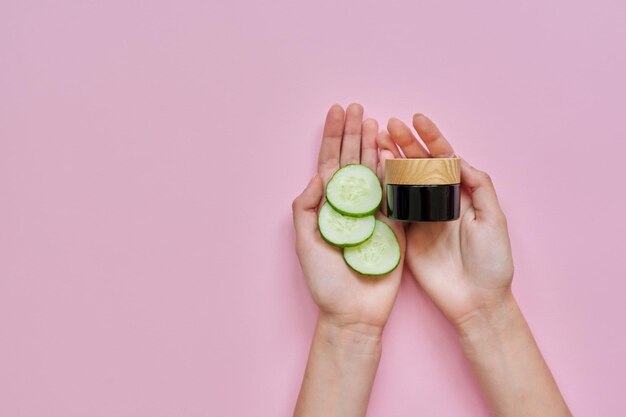 Natural organic cosmetics for face and body woman's hands with glass bottle cucumber slices on pink background copy space