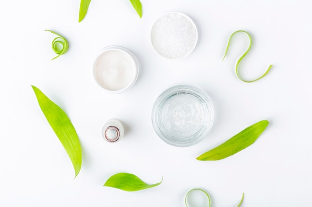 Natural organic homemade cosmetics concept. Skin care, remedy and beauty products: containers with cream and serum among green leaves on white  surface. Flat lay, copy space for text