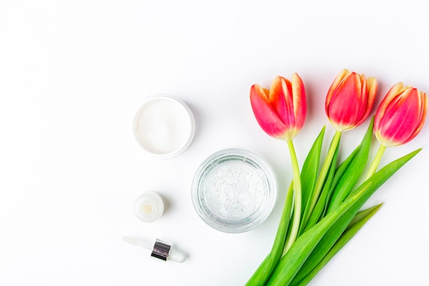 天然オーガニックの自家製化粧品コンセプト。スキンケア、治療、美容製品：白い背景の春の赤いチューリップの花の中でクリームと血清の容器。フラットレイアウト、テキスト用のコピースペース