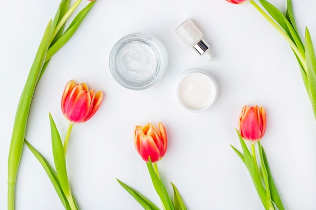 Natural organic homemade cosmetics concept. Skin care, remedy and beauty products: containers with cream and serum among spring red tulip flowers on white surface. Flat lay, copy space for text