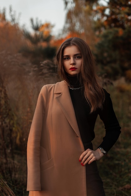 Natural portrait of a beautiful girl with red lips in a fashionable beige coat and a black sweater in the grass on nature. Elegant female style and beauty