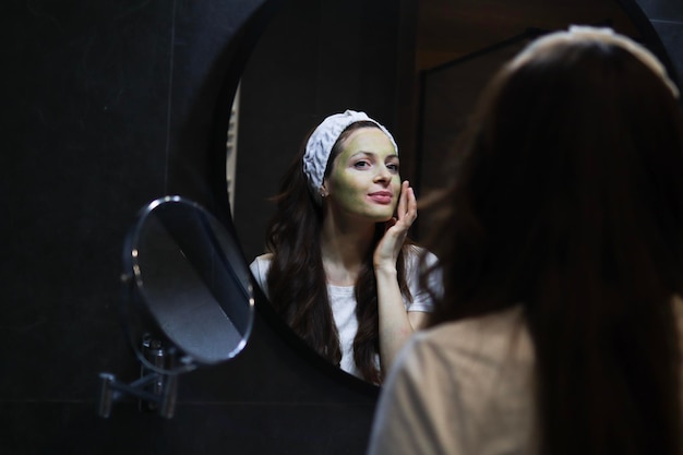 Natural skincare Beauty home spa Woman in headband applying a green clay mask to her face in a modern loft interior bathroom in front of a mirror