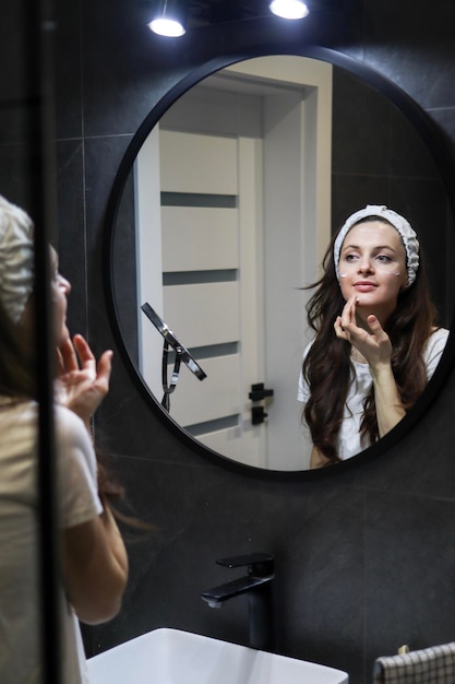 Natural skincare A young woman in a headband making face moistening procedures for her skin in front of a mirror Loft interior bathroom Beauty home spa with vegan cosmetics