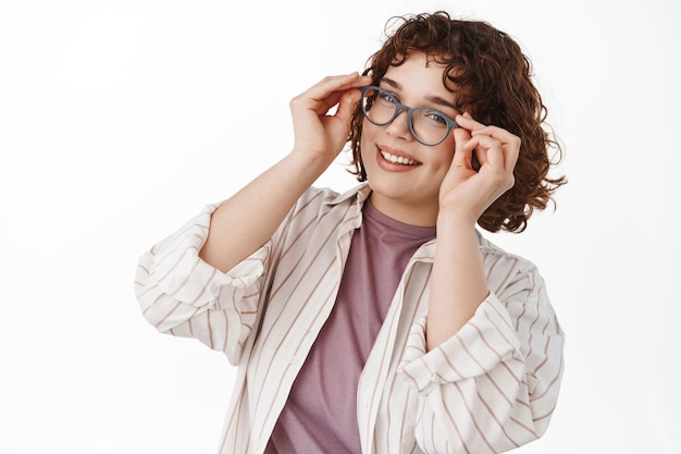 Natural smiling girl with curly hair, put on glasses and looking happy, trying new eyewear at optician store, standing in casual clothes on white.