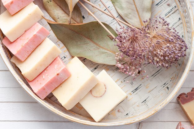 Barra di sapone naturale in un cestino di vimini sopra una priorità bassa di legno bianca e una decorazione secca delle foglie e dei fiori.