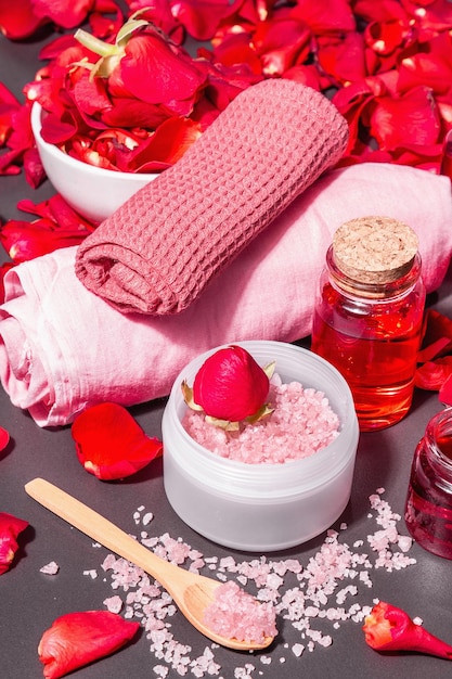 Natural spa concept with fresh roses petals. Rose sea salt, essential oil, soft towels. Modern hard light, dark shadow. Black stone concrete background, copy space