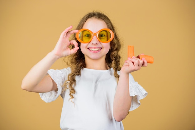 Fonte naturale di vitamine la ragazza mangia la verdura di carota e beve il succo di carota nutrizione vitaminica occhiali da sole per bambini alla moda bere succo vitaminico rinfrescante assistenza sanitaria dieta vitaminica estiva infanzia felice