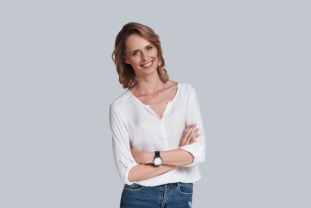 Naturally beautiful. Attractive young woman in casual wear looking at camera and smiling while standing against grey background