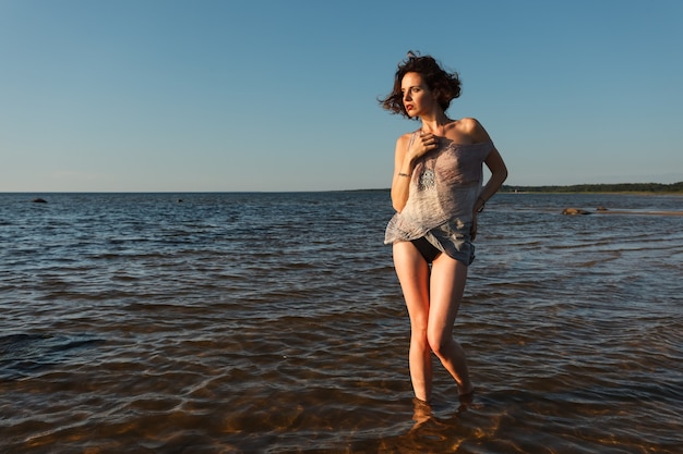 Nature, beauty, youth and healthy lifestyle concept. Attractive young seminude woman in a wet suit posing against the sea background. Evening light with deep shadows and color shift