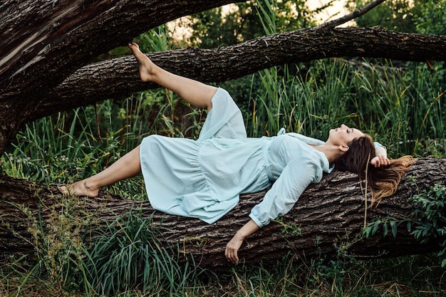 자연과 정신 건강 드레스를 입은 맨발의 여성이 자연의 나무 근처에서 쉬고 있다