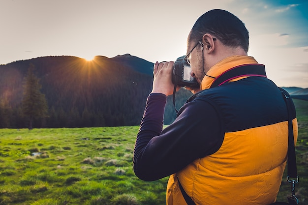 Фото Турист-фотограф природы с камерой, делающей снимок в горах. мечтательный пейзаж заката, весенний зеленый луг и вершина горы в bsckground. вид сзади