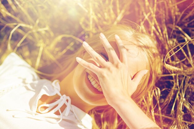 nature, summer holidays, vacation and people concept - close up of happy young woman lying on cereal field and covering face by hand