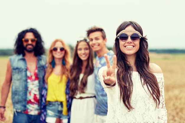 nature, summer, youth culture, gesture and people concept - smiling young hippie friends in sunglasses showing peace hand sign on cereal field