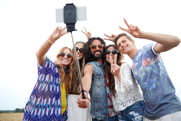 nature, summer, youth culture, technology and people concept - smiling young hippie friends in sunglasses taking picture by smartphone on selfie stick and showing peace gesture on cereal field