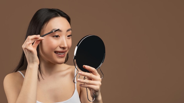 Natuurlijke make-up concept gelukkige aziatische dame die wenkbrauwen borstelt met borstel en spiegel bruine achtergrond