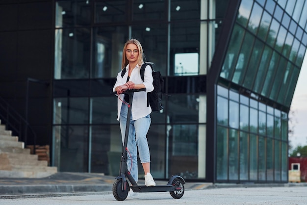 Near business building Beautiful blonde in casual clothes riding electric schooter outdoors at sunny daytime