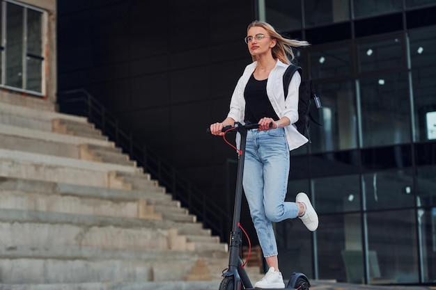 Near business building Beautiful blonde in casual clothes riding electric schooter outdoors at sunny daytime