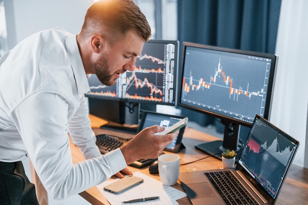 Near the table Young businessman in formal clothes is in office with multiple screens