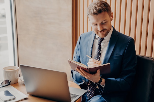 Neat unshaven handsome professional entrepreneur in blue suit holds online financial meeting at laptop with earbuds, sitting by window in cafe with cup of cappuccino, while making notes in workbook