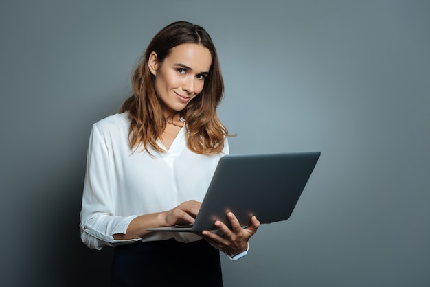 Necessary device. Delighted positive good looking woman holding her laptop and using it for work while standing against grey background