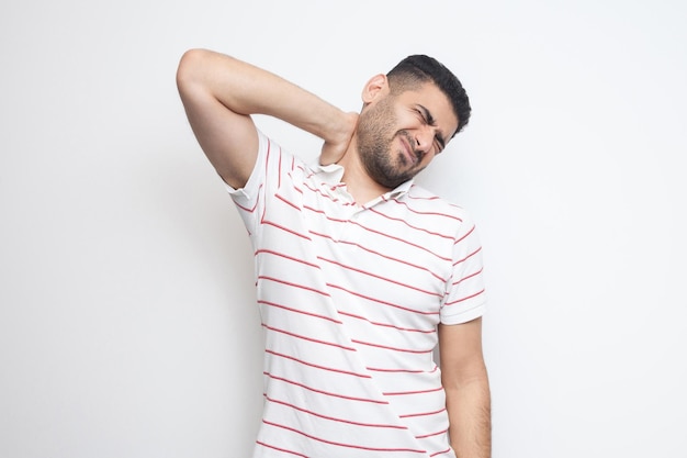 Neck ache or pain. Portrait of handsome bearded young man in striped t-shirt standing and holding his painful neck. indoor studio shot, isolated on white background.