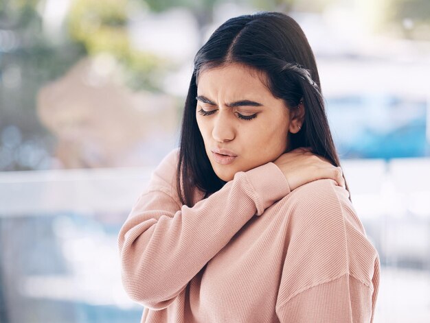 首の痛みと、けいれんに苦しんでいる間、ガラスの背景に彼女の肩を保持しているインドの女性とのモックアップ 医療解剖学または筋肉と、怪我に苦しんでいる魅力的な若い女性