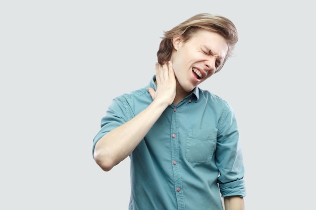 Neck pain. Portrait of sick handsome long haired blonde young man in blue casual shirt standing holding his painful neck because feeling bad. indoor studio shot, isolated on light grey background.