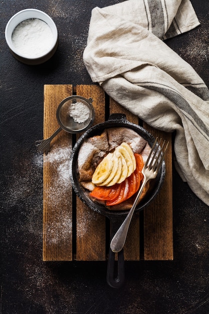Nederlandse baby pannenkoek met appel, kaki, banaan, kaneel in kleine ijzeren pan op houten tafel oppervlak. Bovenaanzicht.